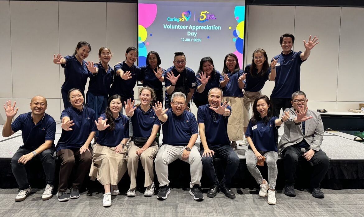 A group of CaringSG team members pose joyfully in two rows, all wearing matching navy blue shirts and holding up five fingers to celebrate CaringSG’s 5th anniversary. They are smiling warmly in front of a screen displaying “Volunteer Appreciation Day – 12 July 2025 – 5 Years & Beyond.” The atmosphere is cheerful and celebratory.